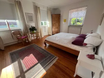 Ein gemütliches Schlafzimmer mit einem großen Bett und bunten Kissen. Die Fenster lassen viel Licht herein und der Holzfußboden sorgt für eine warme Atmosphäre.