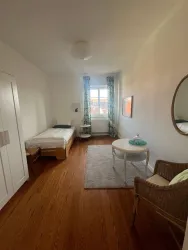 Ein einfaches Schlafzimmer mit einem Einzelbett und einem kleinen Tisch. Die Wände sind weiß, und große Fenster lassen viel Licht herein.