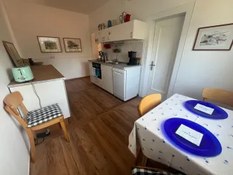 Eine kleine, helle Küche mit einem Tisch und zwei Stühlen. Der Tisch ist mit blauen Tellern und einer weißen Tischdecke gedeckt.