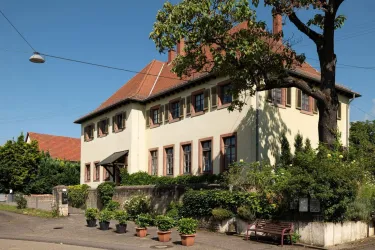 Ein großes, helles Gebäude mit einem roten Satteldach und vielen Fenstern. Umsäumt von grünen Pflanzen und Bäumen bietet es eine einladende Atmosphäre.