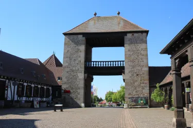 Ein historisches Tor aus Stein steht im Vordergrund. Links und rechts sind traditionelle Gebäude zu sehen, und der Himmel ist klar und blau.