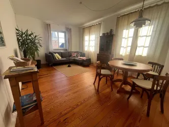 Ein gemütliches Wohnzimmer mit Holzfußboden und großen Fenstern. Es gibt eine Sitzecke, einen Esstisch und eine Pflanze in der Ecke.