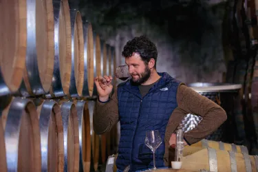 Ein Mann steht in einem Weinkeller und hält ein Glas Wein. Hinter ihm sind große Holzfässer zu sehen.