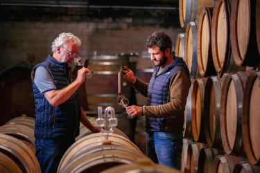 Zwei Männer stehen in einem Weinkeller neben Holzfässern. Sie prüfen den Wein und sprechen miteinander.