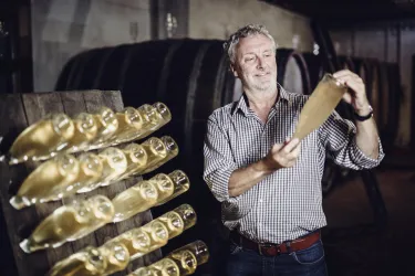 Ein Mann in einem Weinkeller hält eine Flasche in der Hand. Im Hintergrund sind weitere Flaschen auf einem Regal zu sehen.