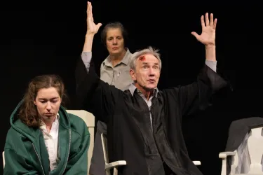 Eine dramatische Theaterszene mit drei Schauspielern. Ein Mann hebt beide Arme, während zwei Frauen aufmerksam zuschauen.