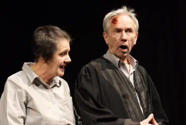 Zwei Schauspieler auf der Bühne in einem emotionalen Moment. Der Mann trägt eine Robe und hat eine Verletzung am Kopf, während die Frau ihn ansprechen scheint.