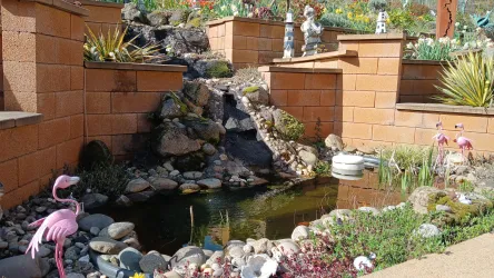 Ein kleiner Teich mit bunten Steinen und Pflanzen umgeben von Ziegelmauern. Auf der linken Seite stehen pinke Flamingo-Dekorationen.