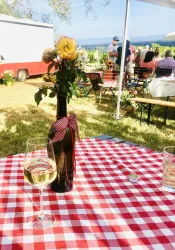 Ein schöner Tisch mit einer rot-weißen Tischdecke, einem Glas Weißwein und einer Blumenvasen. Im Hintergrund sind Menschen und eine malerische Landschaft zu sehen.