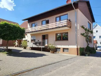 Ein einladendes Wohnhaus mit einem Balkon und einem gepflasterten Vorhof. Der Himmel ist klar und blau, was eine angenehme Atmosphäre schafft.