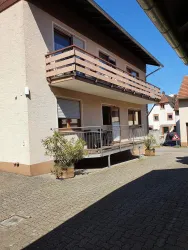 Ein zweigeschossiges Wohnhaus mit einem Balkon und einer Terrasse. Der Himmel ist klar und das Wetter scheint sonnig zu sein.