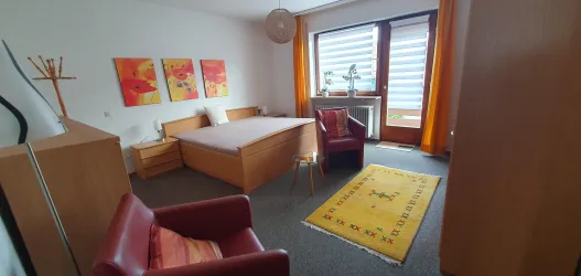Ein helles Schlafzimmer mit einem großen Bett und bunten Bildern an der Wand. Ein roter Stuhl und ein Teppich vervollständigen den Raum.