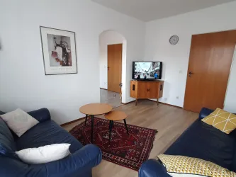 Ein gemütliches Wohnzimmer mit zwei blauen Sofas und einem Holztisch. An der Wand hängt ein Bild und ein Fernseher zeigt ein Programm.