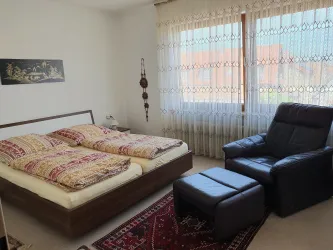 Ein gemütliches Schlafzimmer mit einem Doppelbett und bunten Decken. Im Raum steht ein schwarzer Sessel und ein kleiner Hocker.