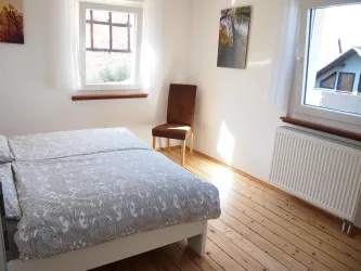 Ein helles Schlafzimmer mit einem Doppelbett und einem Stuhl. Große Fenster sorgen für viel Tageslicht.