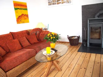 Ein gemütliches Wohnzimmer mit einer orangefarbenen Couch und einem Glastisch. Auf dem Tisch stehen eine gelbe Kerze und ein Blumengesteck.