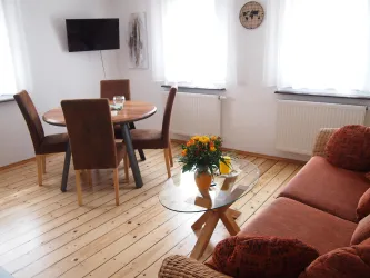 Ein gemütlicher Wohnraum mit einem runden Esstisch und braunen Stühlen. Auf dem Tisch steht eine Blumenvase, und ein Sofa ergänzt die Einrichtung.