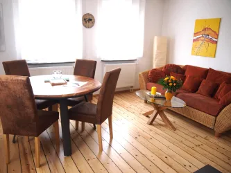 Ein gemütliches Wohnzimmer mit einem runden Holztisch und vier Stühlen. Auf der Couch liegen rote Kissen, und ein Tisch mit einer Blumenvase und einer Kerze steht in der Mitte.