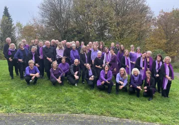 Eine große Gruppe von singen Menschen steht im Freien. Sie tragen lila Kleidung und lächeln freundlich in die Kamera.