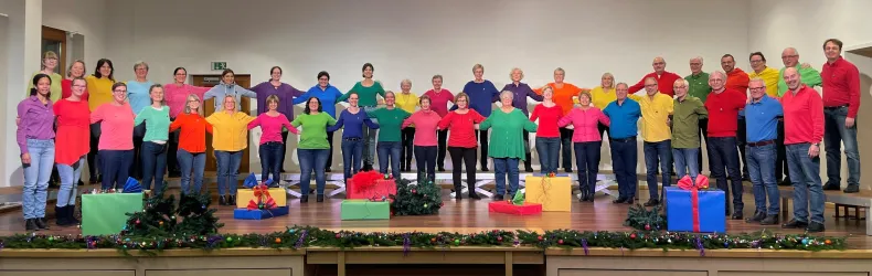 Eine große Gruppe von Menschen steht in bunten Oberteilen auf einer Bühne. Vor ihnen befinden sich Geschenke in verschiedenen Farben.
