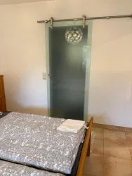 Ein schlichtes Schlafzimmer mit einem Doppelbett und gefalteten Handtüchern. Eine gläserne Schiebetür führt zu einem anderen Raum.