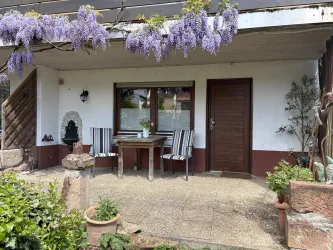Eine einladende Terrasse mit einem Tisch und zwei Stühlen. Über ihr blühen lila Wisteria-Blüten und grüne Pflanzen verleihen dem Raum einen dekorativen Touch.