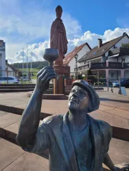 Eine Bronzestatue eines Mannes, der ein Glas hält, steht im Vordergrund. Im Hintergrund sieht man eine weitere Statue sowie mehrere Gebäude und einen blauen Himmel.