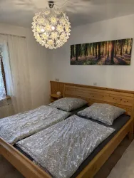 Ein gemütliches Schlafzimmer mit einem Holzbett und eleganter Bettwäsche. An der Wand hängt ein Bild mit einem Waldmotiv und eine stilvolle Deckenleuchte sorgt für angenehmes Licht.