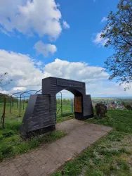 Ein architektonisches Tor mit der Aufschrift "Deutsches Weintor" in einer grünen Landschaft. Der Himmel ist teils bewölkt und blau.