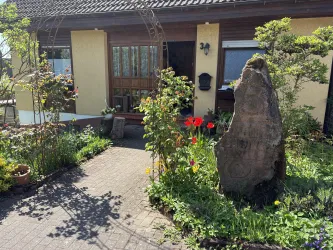 Ein gemütliches Haus mit einem einladenden Eingang und bunten Blumen im Garten. Ein markanter Stein neben dem Weg ergänzt die idyllische Atmosphäre.