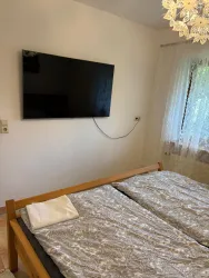 Ein Schlafzimmer mit einem großen Bett und einem Fernseher an der Wand. Durch die Fenster fällt sanftes Licht auf die Bettwäsche.