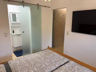 Ein modernes Schlafzimmer mit einem Doppelbett und einem Fernseher an der Wand. Im Hintergrund ist ein Badezimmer durch eine Glaswand sichtbar.