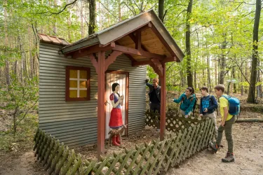 Eine kleine Hütte im Wald mit einer Person in einem roten Kleid. Mehrere Kinder beobachten neugierig von außen.