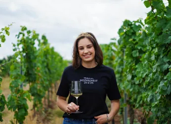 Eine Frau in einem Weingarten hält ein Glas Wein in der Hand. Sie trägt ein schwarzes T-Shirt und lächelt vor einer Reihe von Weinreben.