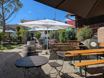 Ein einladender Außenbereich mit Holztischen und Sitzbänken unter Sonnenschirmen. Im Hintergrund sind grüne Pflanzen und ein rustikales Gebäude zu sehen.