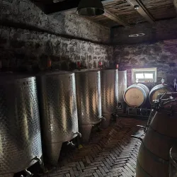 Ein Innenraum einer Weinlagerung mit großen, silbernen Tanks und Holzfässern. Die Wände sind aus Stein und es gibt ein Fenster, das Tageslicht hineinlässt.