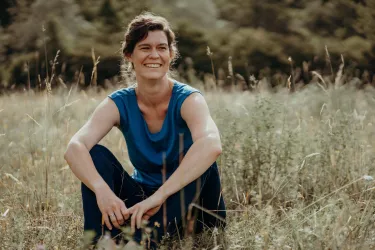 Eine lächelnde Frau sitzt im Gras und genießt die Natur. Sie trägt ein blaues T-Shirt und wirkt entspannt.
