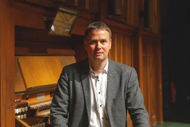 Ein Mann steht vor einer Orgel in einem Bühnenraum. Er trägt einen grauen Blazer und ein weißes Hemd.