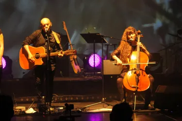 Ein Musiker singt und spielt Gitarre, während eine Cellistin neben ihm spielt. Hintergrundlich sind bunte Lichter zu sehen, die eine lebendige Atmosphäre schaffen.