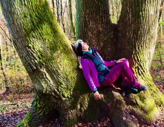 Eine Person lehnt entspannt an einem großen Baum in einem Wald. Die Umgebung ist herbstlich mit buntem Laub.
