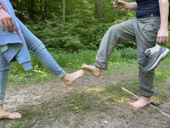 Zwei Personen stehen barfuß auf einem Waldweg und berühren mit den Füßen einander. Der Hintergrund zeigt grüne Bäume und eine natürliche Umgebung.