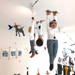 Eine bunte mobile Skulptur mit Figuren, die an Stangen hängen. Im Hintergrund sind weitere künstlerische Elemente und Bilder zu sehen.