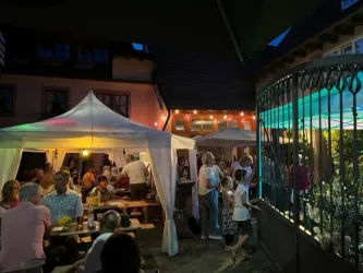 Eine lebhafte Sommernacht mit vielen Menschen, die in einem gemütlichen Garten feiern. Zelte und stimmungsvolle Beleuchtung sorgen für eine einladende Atmosphäre.
