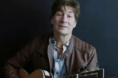 Ein Mann mit einer Gitarre lächelt in die Kamera. Er trägt eine Lederjacke und sitzt vor einem dunklen Hintergrund.