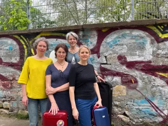 Vier Frauen stehen vor einer bunten Graffiti-Wand. Sie halten Koffer und blicken lächelnd in die Kamera.