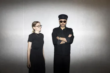 Ein Musiker singt und spielt Gitarre, während eine Cellistin neben ihm spielt. Hintergrundlich sind bunte Lichter zu sehen, die eine lebendige Atmosphäre schaffen.