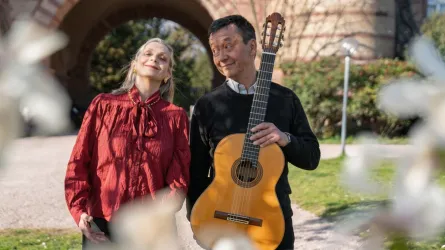 Ein Mann hält eine Gitarre und lächelt, während eine Frau neben ihm steht. Im Hintergrund sind Blumen und ein schöner Park zu sehen.
