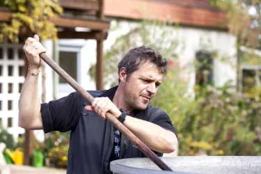 Ein Mann arbeitet mit einem Besenstiel in einem Garten. Im Hintergrund sind Pflanzen und ein Haus zu sehen.