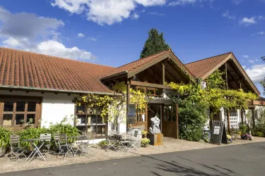 Ein charmantes Gebäude im ländlichen Stil mit einem roten Ziegeldach und schönen grünen Pflanzen. Draußen stehen einige Tische und Stühle für Gäste.