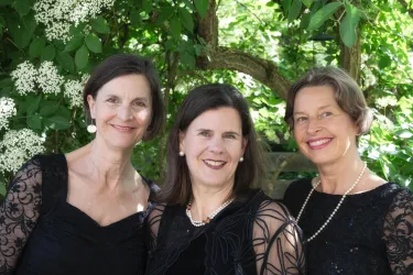 Drei Frauen in eleganten schwarzen Kleidern stehen lächelnd vor einem Hintergrund mit blühenden Pflanzen. Sie scheinen einen festlichen Anlass zu genießen.
